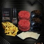 Halloween-Burger-Bundle mit zwei Rindfleisch-Pattys, zwei schwarzen Burgerbrötchen mit Sesam, Käsescheiben, Pommes frites, BBQ-Sauce und Gewürzmischung auf dunklem Hintergrund mit Halloween-Dekoration.