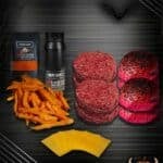 Halloween-Burger-Bundle-Rot mit zwei rohen Rindfleisch-Pattys, zwei roten Burgerbrötchen mit schwarzem Sesam, Süßkartoffel-Pommes, Käsescheiben und zwei Gewürzpackungen. Halloween-Dekoration mit Fledermaus und Kürbis im Hintergrund.